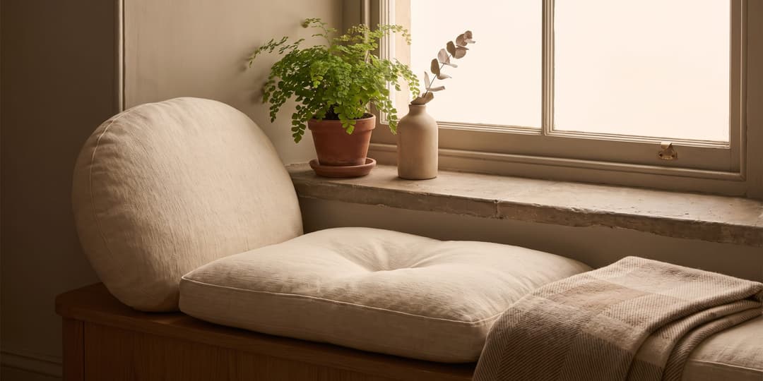 A cushioned window seat with a round bolster cushion, a potted fern and a ceramic vase with eucalyptus on a stone windowsill, and a folded plaid blanket.