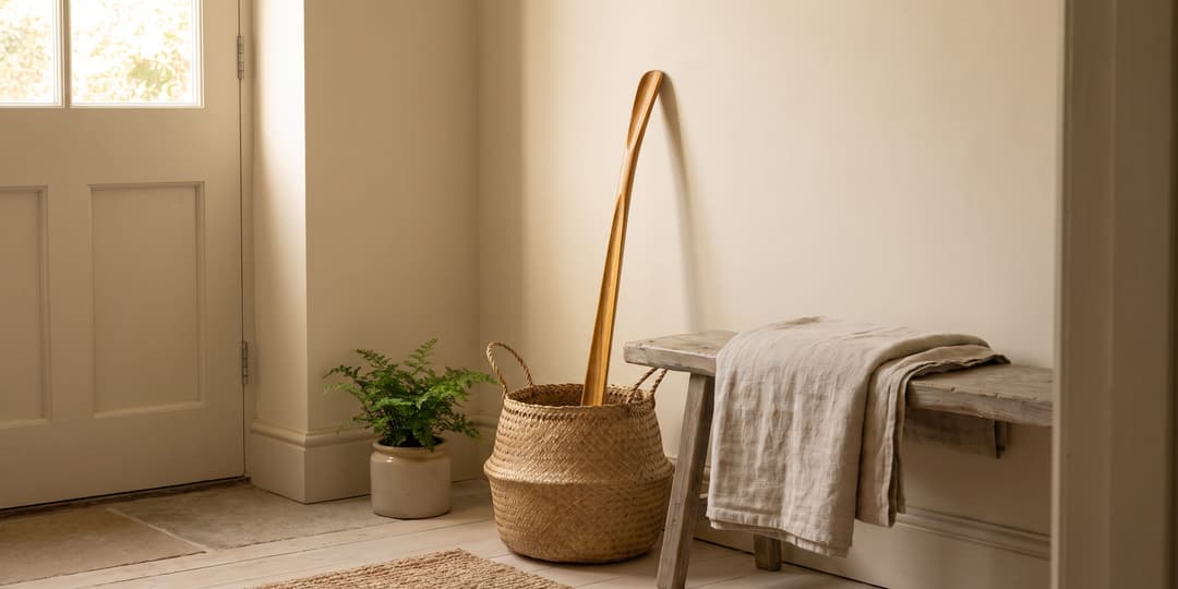 A woven basket holding a long curved wooden handle sits beside a rustic wooden bench draped with a linen throw near a potted fern and a white paneled door.