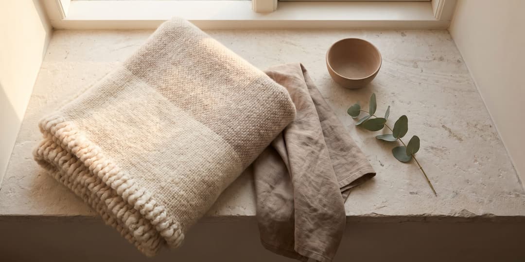 A folded wool blanket and linen cloth beside a small wooden bowl and a eucalyptus sprig on a stone windowsill.