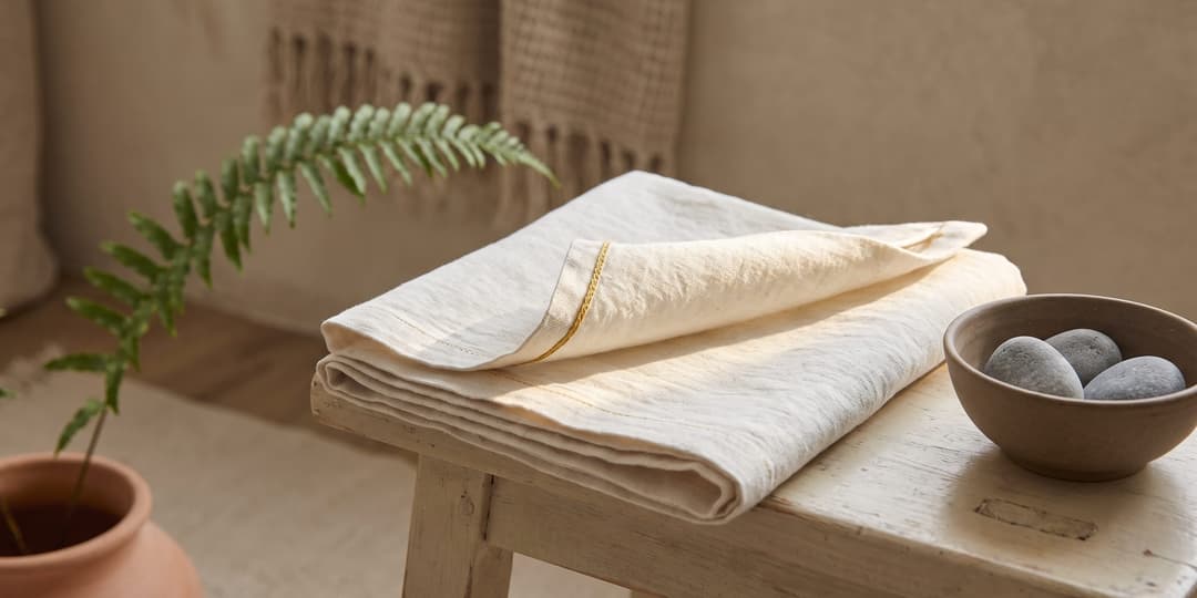 A folded piece of linen rests on a wooden stool with one corner peeled back, beside a small ceramic bowl of stones and a fern in a terracotta pot, lit by soft natural light.