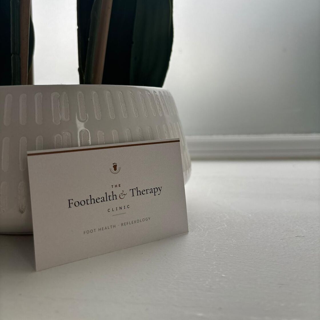 A Foothealth and Therapy Clinic business card propped against a ribbed white plant pot on a windowsill