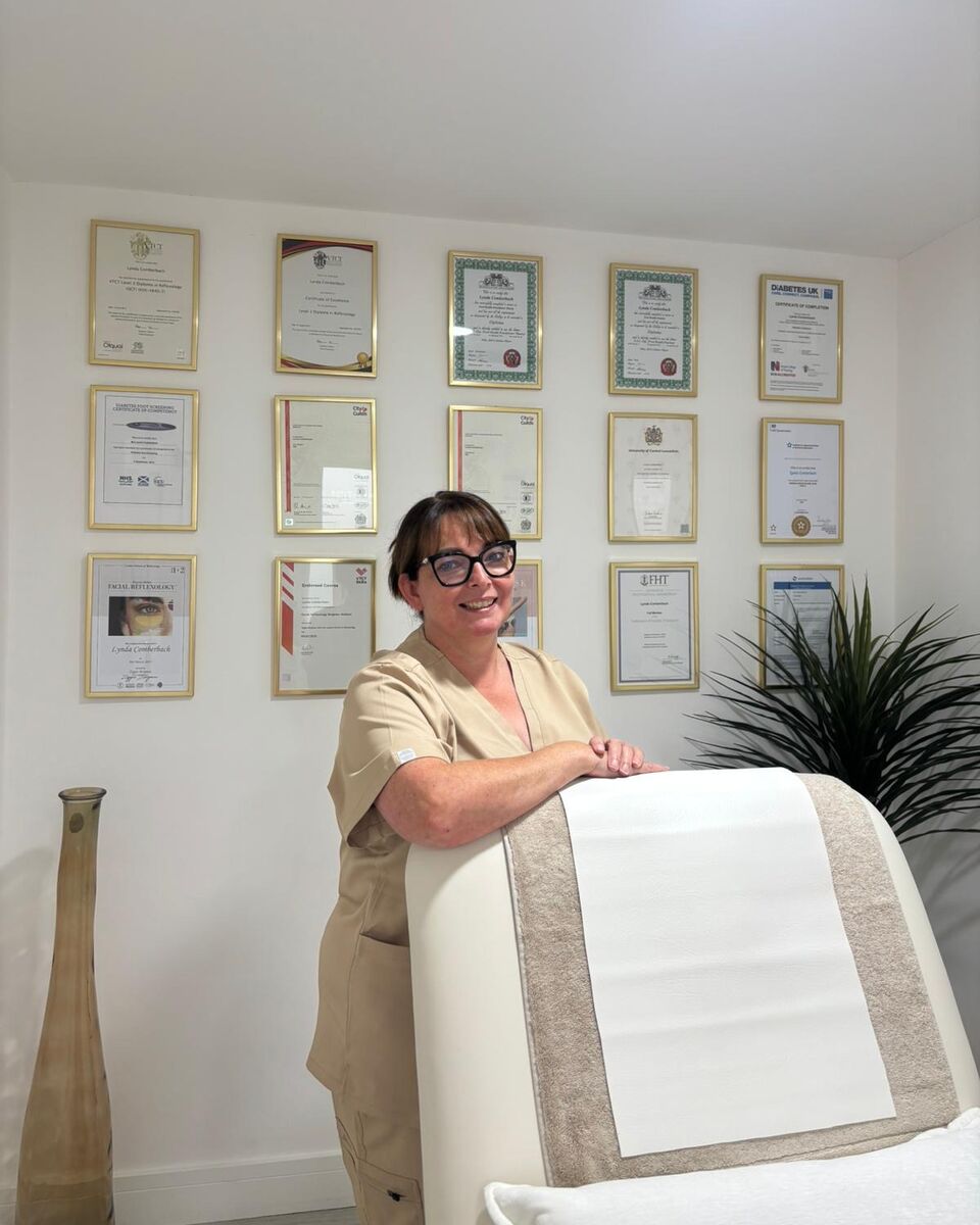 Lynda Comberbach in her beige clinic uniform, standing beside the treatment couch in front of the wall of framed qualifications in her Hapton clinic