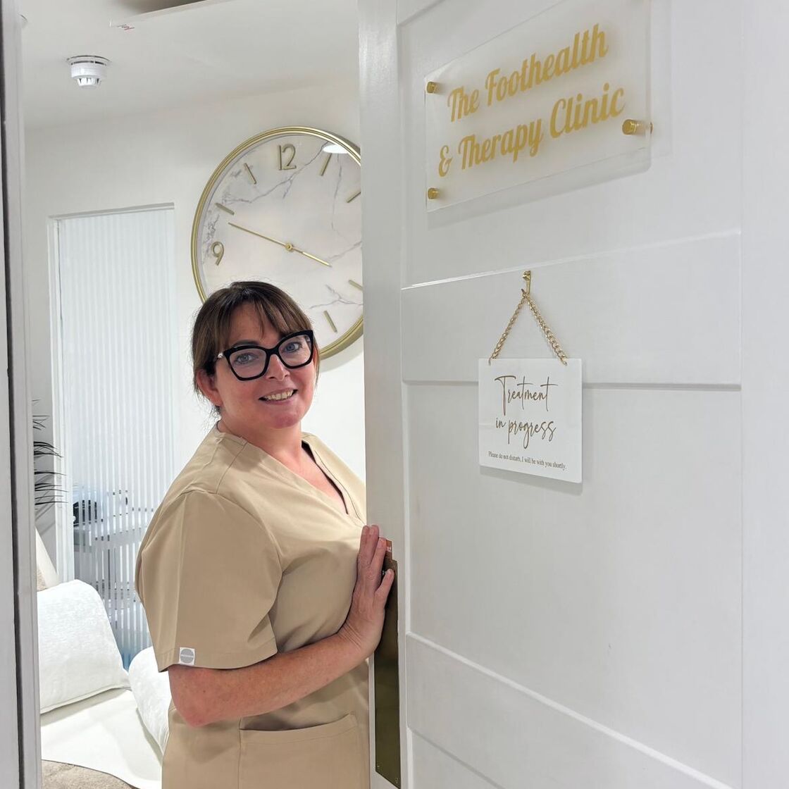 Lynda Comberbach in her beige clinic uniform, holding the clinic door open beneath the gold clinic sign and a Treatment in progress card