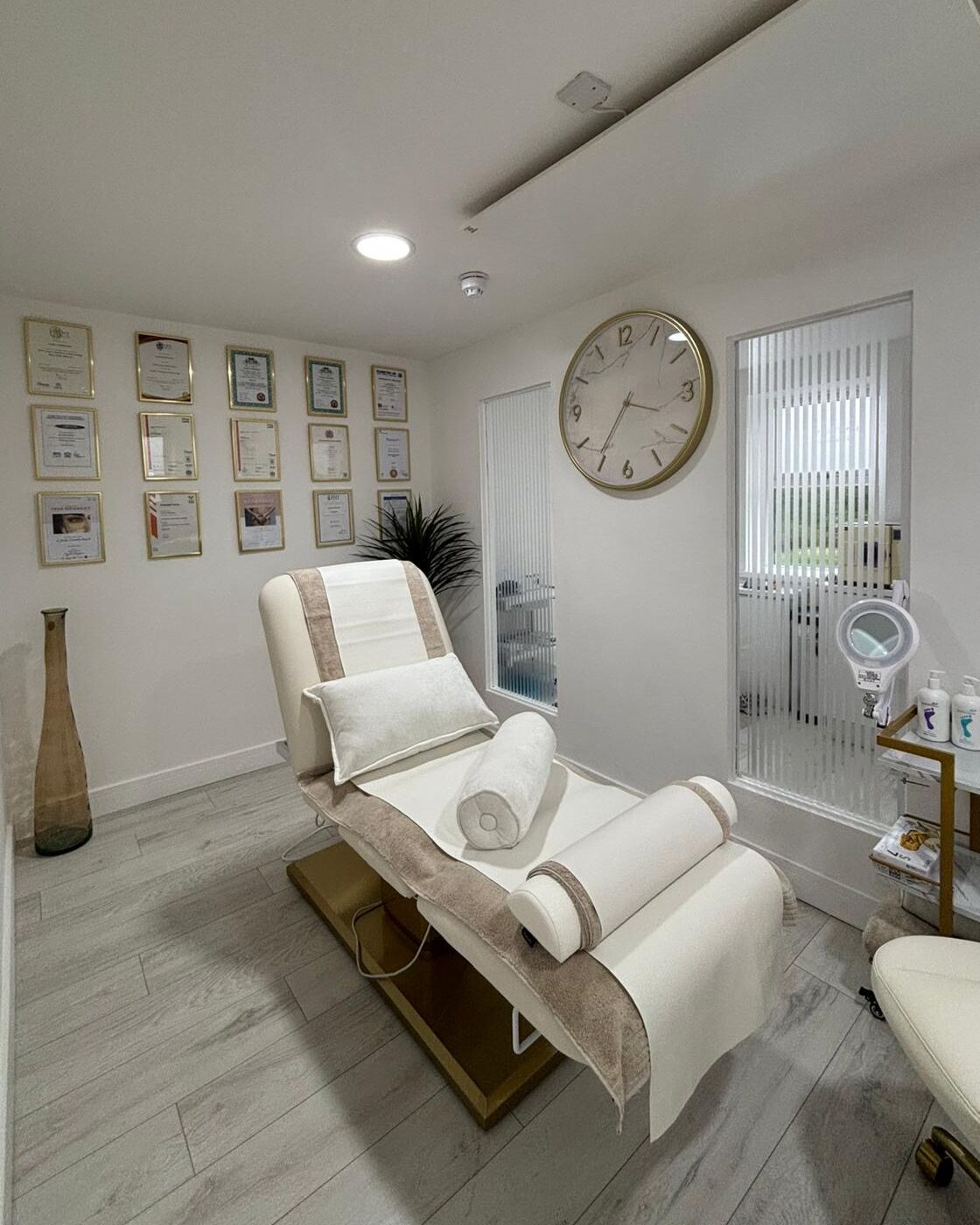 The treatment room at the clinic in Hapton, with a cream reclining treatment couch and bolster cushion, a wall of framed qualifications, a large gold rimmed wall clock and pale wood effect flooring