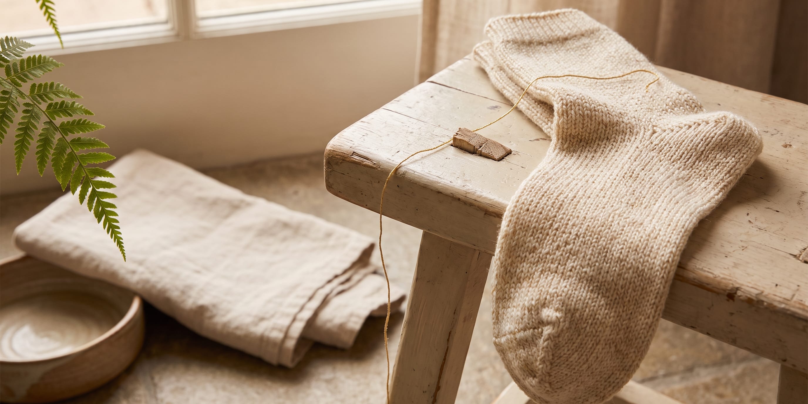 A pair of hand-knitted cream wool socks draped over a distressed wooden bench beside a spool of gold thread, with folded linen, a ceramic bowl, and a fern in the background near a window.