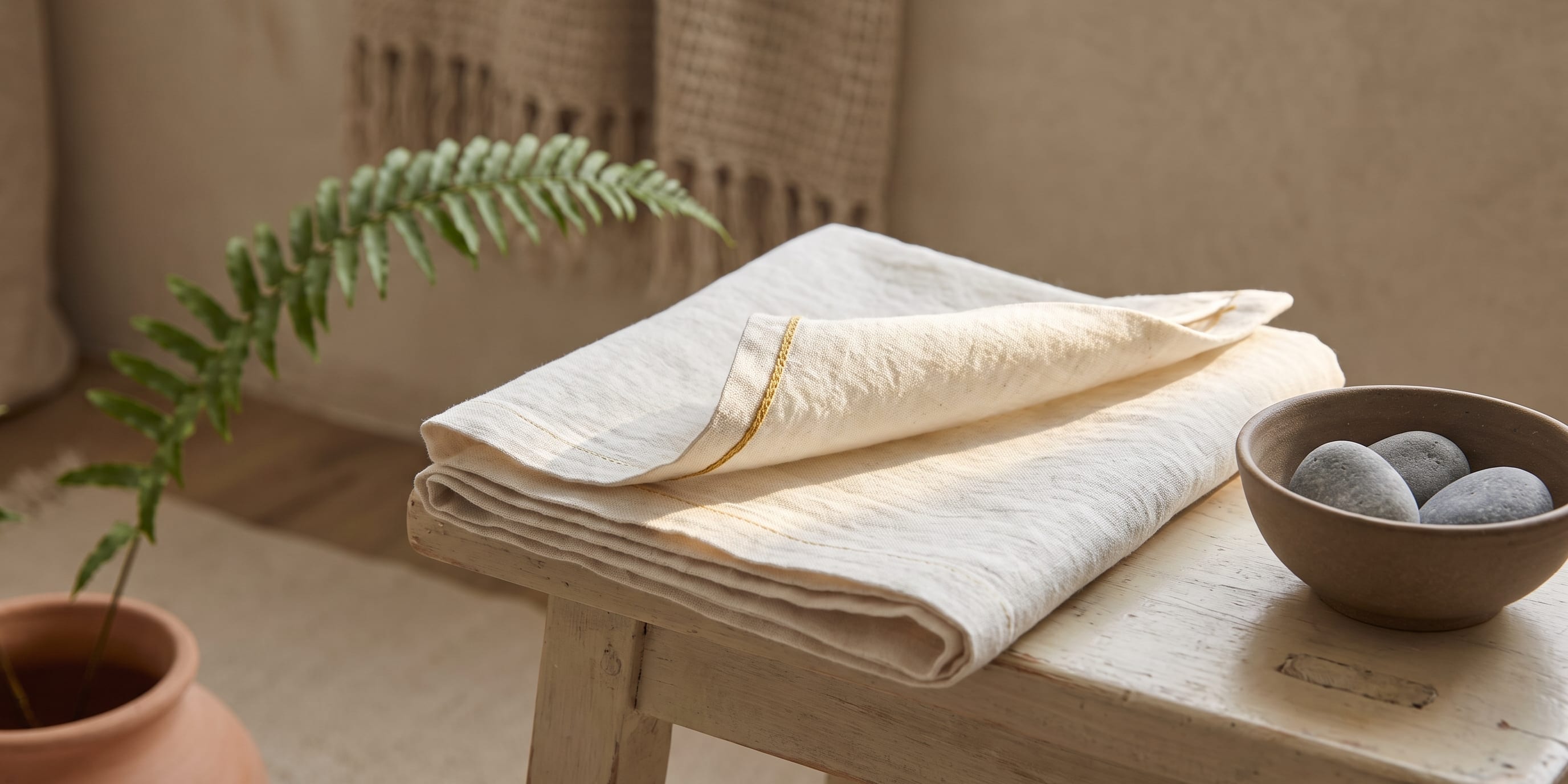A folded piece of linen rests on a wooden stool with one corner peeled back, beside a small ceramic bowl of stones and a fern in a terracotta pot, lit by soft natural light.